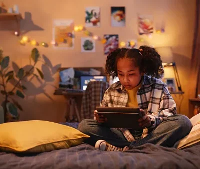 teenage black girl using digital tablet sitting on bed at home in warm light